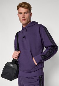 Hoodie roxo feito de uma mistura de algodão com riscas pretas nas mangas e um bolso frontal. O modelo segura uma pequena bolsa preta com uma alça.