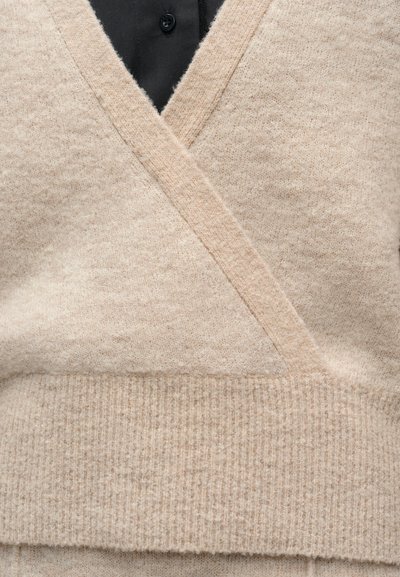 Μπεζ πλεκτή ζακέτα με ριμπ τελείωμα, φορεμένη πάνω από μαύρο πουκάμισο με κουμπιά.