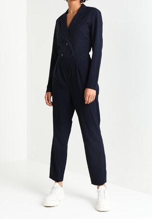 Kobieta ubrana w granatowy, długorękawowy, dopasowany kombinezon z dwurzędowymi guzikami i białe masywne trampki, stojąca na białym tle.