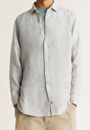 Mann mit einem hellgrau-weiß vertikal gestreiften Hemd über einem blauen T-Shirt und beigefarbenen Hosen, die Hände in den Taschen, neutraler Hintergrund.