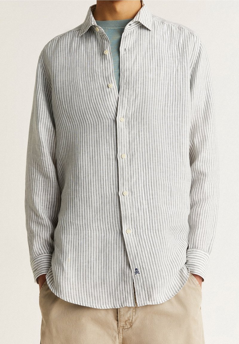Homme portant une chemise à rayures verticales gris clair et blanc sur un t-shirt bleu avec un pantalon beige, les mains dans les poches, fond neutre.