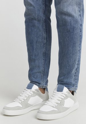 White sneakers with grey suede accents and light blue heel tabs. The design features a textured surface and a flat rubber sole, paired with blue jeans.