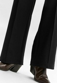 Pantaloni neri a gamba larga realizzati in tessuto liscio, con orlo pulito. Scarpe con tacco marroni in pelle testurizzata e punta quadra.