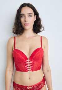 Sutien balconette din dantelă roșie și satin, cu decolteu adânc, panouri transparente și detalii decorative cu șireturi în față. Chiloți asortați din dantelă.