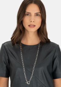 Sølv kædeled med forlængede led og en cirkulær lås, båret over en sort lædertop med geometriske sømdetaljer.