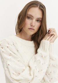 Weißer Strickpullover mit lockerer Passform, texturiertem Muster und gerippten Bündchen. Das Modell hält die Hände nah an ihrem Gesicht und zeigt weiche Fasern.