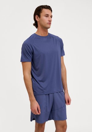 Mann steht in einem passenden blauen Sportoutfit mit kurzärmeligem Hemd und Shorts vor einem weißen Hintergrund.