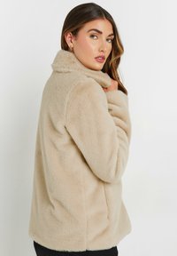 Beige faux fur jakke med høj krave, blød tekstur, afslappet pasform og lange ærmer, designet med en enkelt knaplukning ved halsen.