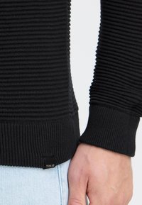 Gros plan sur la main d’une personne portant un poignet de pull noir côtelé et un jean bleu clair, montrant la texture et une petite étiquette de marque.