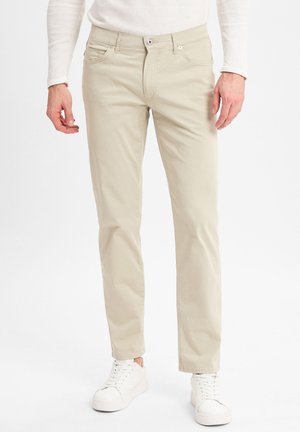 Beige Baumwollhose mit geradem Schnitt, ausgestattet mit Fronttaschen und Reißverschluss, kombiniert mit weißen Sneakern. Einfaches, klares Design.