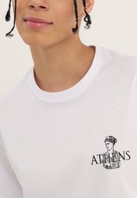 Jongere met gevlochten haar draagt een wit T-shirt met een zwarte busteillustratie en het woord "ATHENE" op de borst.