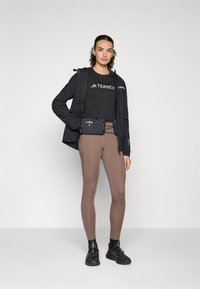 Kvinne står iført svart Terrex-jakke og t-skjorte, brune leggings, svarte joggesko og en svart hofteveske, mot en ensfarget bakgrunn.