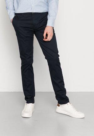 Mann trägt schwarze Slim-Fit-Hose, weiße Sneakers und ein hellblau gestreiftes Hemd, eine Hand in der Tasche.