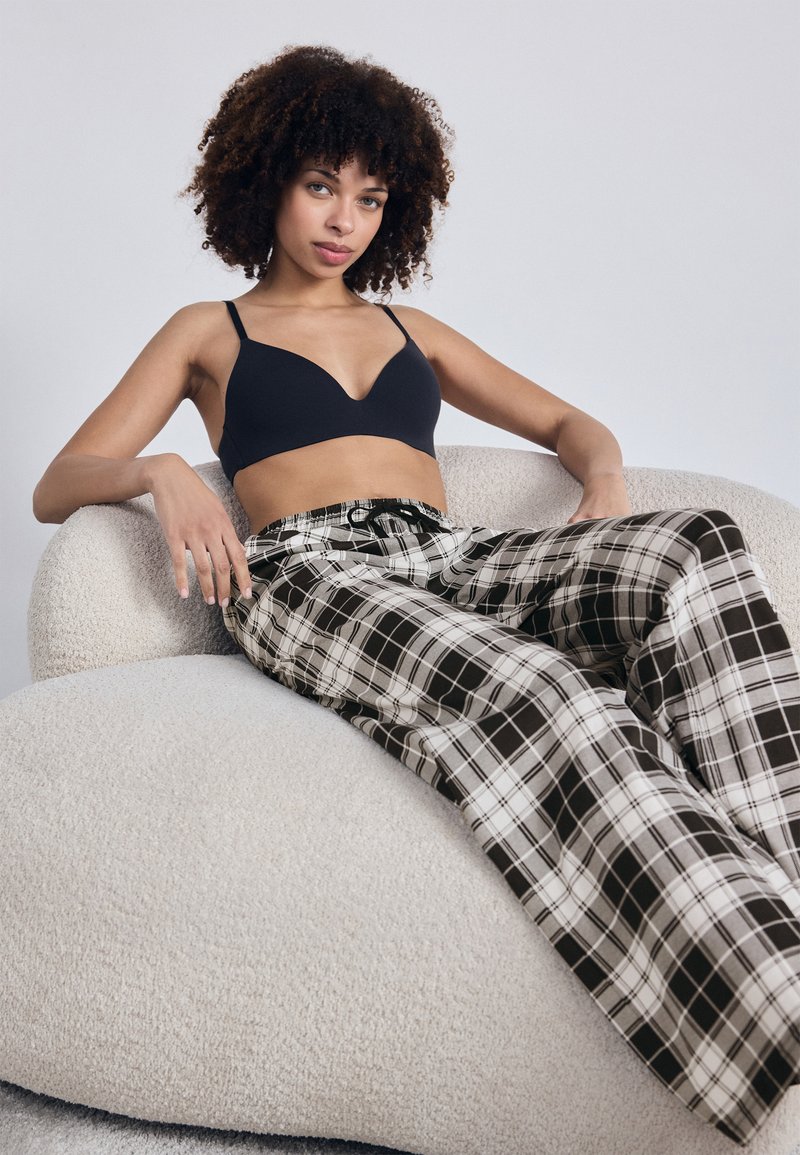 Bralette preta com alças finas, combinada com calças largas em xadrez preto e branco. Modelo sentado numa cadeira texturizada bege.