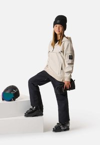 Un giovane indossa una felpa con cappuccio color crema e pantaloni da neve neri, tenendo dei guanti neri. Un casco nero con una visiera oscurata è su una pedana bianca.