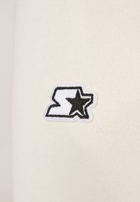 Insignia bordada que presenta una letra 'S' estilizada con una estrella, contorneada en negro sobre un fondo de tela color crema.