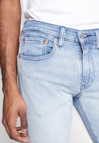 Pantalones vaqueros de mezclilla azul claro con un diseño ajustado, que cuentan con remaches de color cobre, múltiples bolsillos y una textura suave.