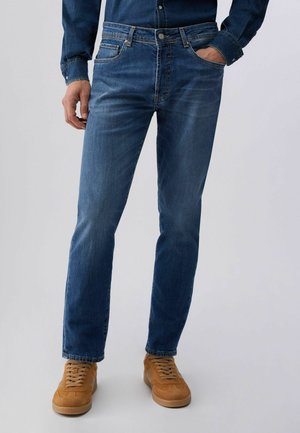 Uomo che indossa jeans blu aderenti, una camicia di denim scuro e scarpe da ginnastica marrone chiaro con lacci, in piedi con una mano in tasca.