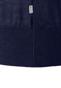 Zbliżenie na granatowy dzianinowy materiał z ribbowanym wykończeniem i małą białą metką oznaczoną "MAERZ", uszytą blisko szwu.