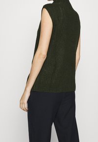 Zelený pletený svetřík bez rukávů s vysokým límcem, zdobený strukturovaným vzorem a s volným střihem, kombinovaný s tmavými žebrovanými kalhotami.