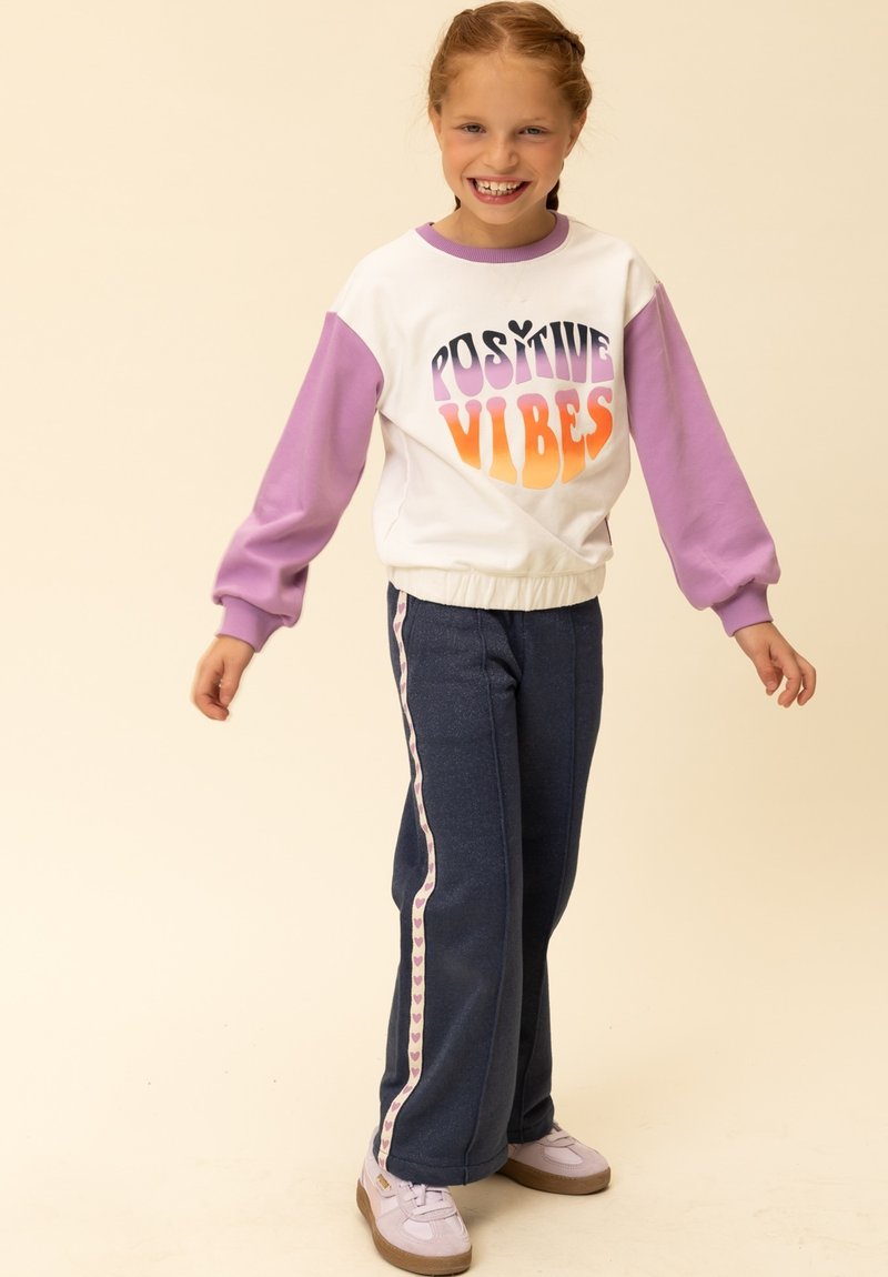 Witte sweater met paarse mouwen en de tekst "POSITIVE VIBES" erop. Blauwe joggingbroek met zijpaneel in hartjespatroon. Lichtpaarse sneakers.