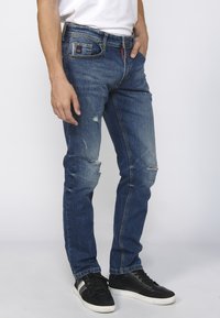 Jeans de mezclilla en azul oscuro con áreas desgastadas y rasguños en las rodillas, combinados con zapatillas negras y una camiseta blanca básica.