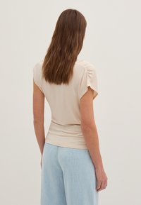 Beige åtsittande topp med korta ärmar och gathered detaljering vid axlarna, matchad med ljusblå vida jeans. Slät textur, minimalistisk design.