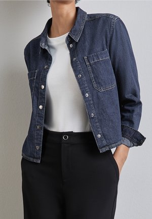 Person iført en mørkeblå denimjakke over en hvid t-shirt og sorte højtaljede bukser med hænderne i lommerne, foran en ensfarvet væg.