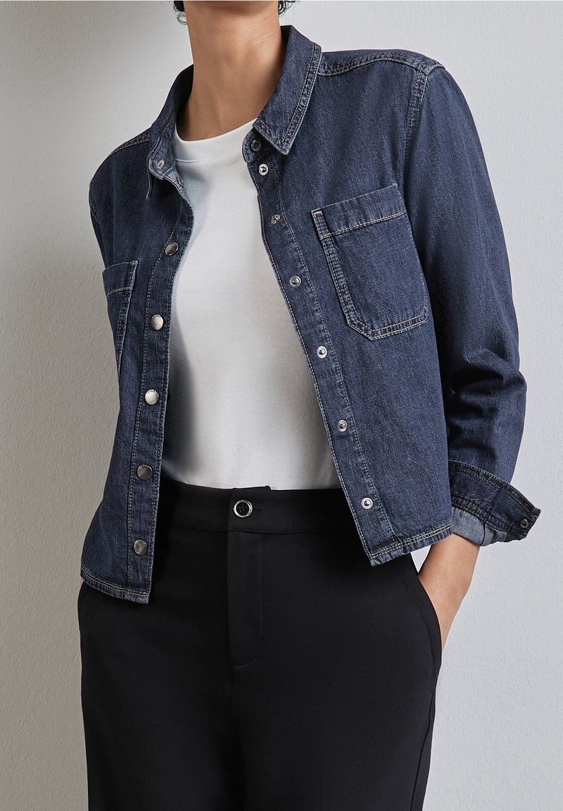 Personne portant une veste en jean bleu foncé par-dessus un t-shirt blanc et un pantalon noir taille haute, les mains dans les poches, devant un mur uni.