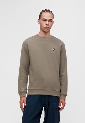 Jeune homme aux cheveux tressés portant un sweat-shirt uni vert olive avec un petit logo jaune sur la poitrine et un pantalon bleu foncé.