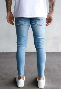 Gai zila skinny džinsi no denima, ar diviem aizmugurējiem kabatām un gludu tekstūru, apvienoti ar baltiem kedām uz pelēka grīdas.