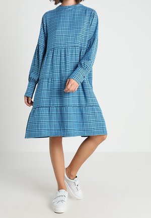 Person wearing a blue plaid long-sleeve knee-length dress with white sneakers, standing against a plain light background.