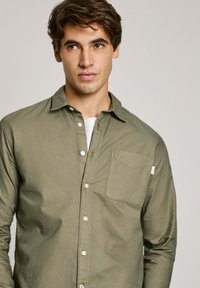 Camicia a maniche lunghe verde oliva in cotone, con taschino sul petto, bottoni bianchi e colletto classico. Tessuto morbido.