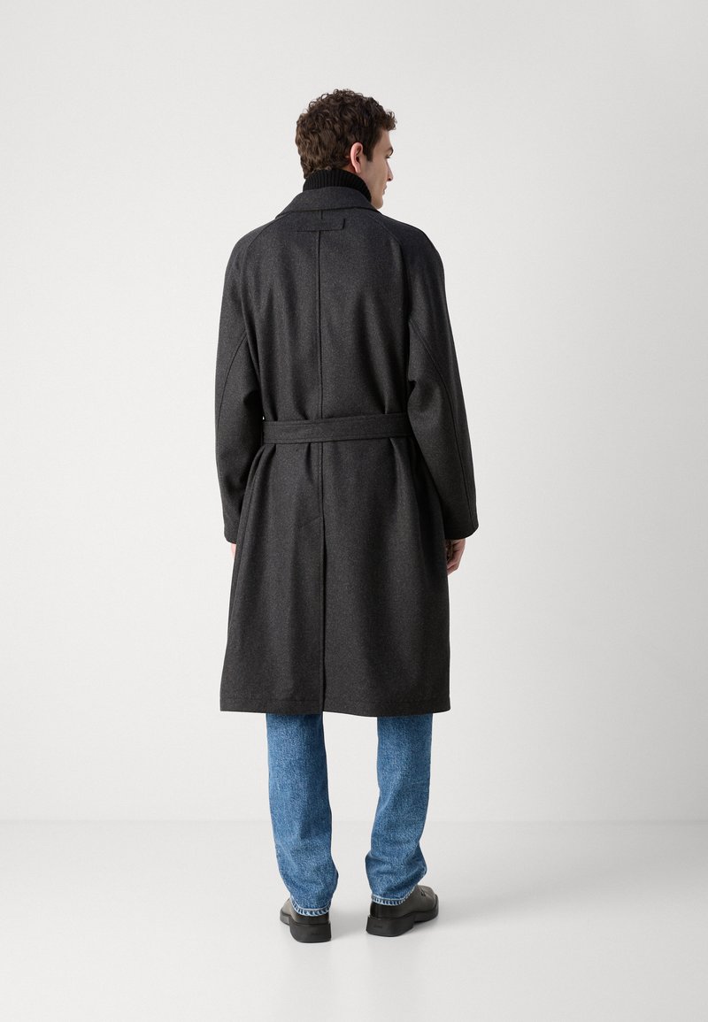 Manteau en laine gris foncé avec une taille ceinturée, des revers larges et une fente au dos, associé à un jean en denim bleu et des chaussures noires.