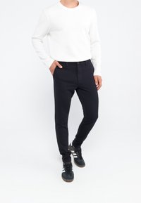 Zwarte slim-fit broek met een gladde textuur, knoopsluiting en voorzakken, gecombineerd met een wit overhemd met lange mouwen en donkere sneakers.