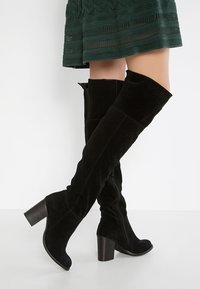Bottes noires au-dessus du genou en suede doux, avec un talon épais et une fermeture éclair intérieure pour un enfilage facile. Tissu texturé avec une finition lisse.