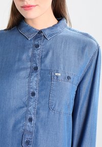 Blå denimskjorta med knäppning fram, spetsig krage, långa ärmar och en liten bröstficka. Tyget har en slät textur.