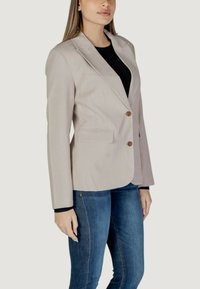 Ανοιχτό μπεζ σακάκι με γιακά με εσοχή και δύο καφέ κουμπιά. Διαθέτει μπροστινές τσέπες και λείο, υφασματοποιημένο υλικό. Φοράει από πάνω μαύρο μπλουζάκι.