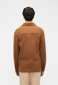 Καφέ μπουφάν πουκάμισο από φλις με γιακά, μανσέτες με κουμπιά και καμπύλο τελείωμα, με υφασμάτινη υφή και ευθεία σιλουέτα.