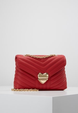 Sac à main matelassé rouge avec surpiqûres en chevrons, fermoir doré en forme de cœur et bandoulière en chaîne dorée, présenté sur un socle blanc.