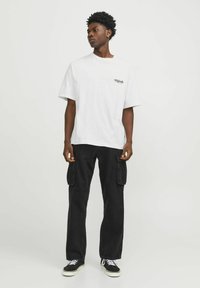 T-shirt bianca in cotone con maniche corte e una piccola stampa; pantaloni cargo neri con molte tasche; sneakers nere con suole bianche.