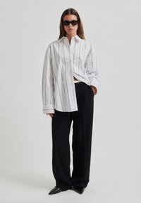 White button-up shirt with black vertical stripes, long sleeves, and a relaxed fit, paired with black wide-leg trousers and pointed black shoes.