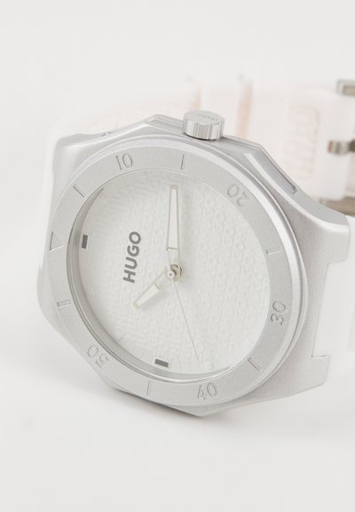 Montre argentée avec un cadran blanc texturé, index heures noirs et un bracelet en silicone blanc. Présente un boîtier rond et des accents en acier inoxydable.