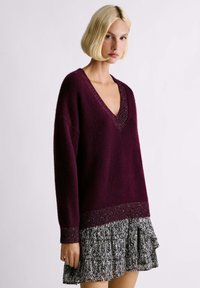 Donna bionda che indossa un maglione a V profondo di colore viola scuro con rifiniture metalliche e una gonna a pieghe con motivo in bianco e nero, in piedi davanti a uno sfondo neutro.