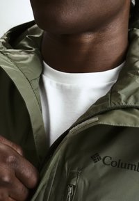Mann trägt olivgrüne Columbia-Jacke über weißem Hemd, öffnet die Jacke teilweise im Brustbereich.