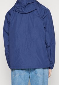 Giacca a vento blu navy realizzata in tessuto testurizzato, con cappuccio regolabile e polsini elasticizzati. Indossata sopra jeans di denim azzurri.