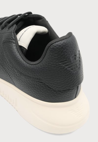 Basket en cuir noir texturé avec semelle blanche, présentant un logo en relief au talon et un détail de col rembourré sur fond lumineux.