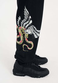 Schwarze Jogginghosen mit einem bunten Grafikdesign von einem Adler und einer Schlange, plus roten Akzenten. Kombiniert mit schwarzen Sneakern mit Luftpolsterung.