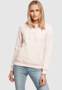 Felpa di cotone rosa chiaro con tasca frontale, cappuccio con cordini e piccolo dettaglio testuale sul petto. Abbinata a jeans blu chiaro.