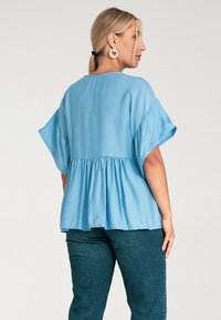 Jasnoniebieska bluzka z krótkim rękawem, z zebranym pasem peplum i gładką fakturą. Połączona z ciemnozielonymi, dopasowanymi spodniami.